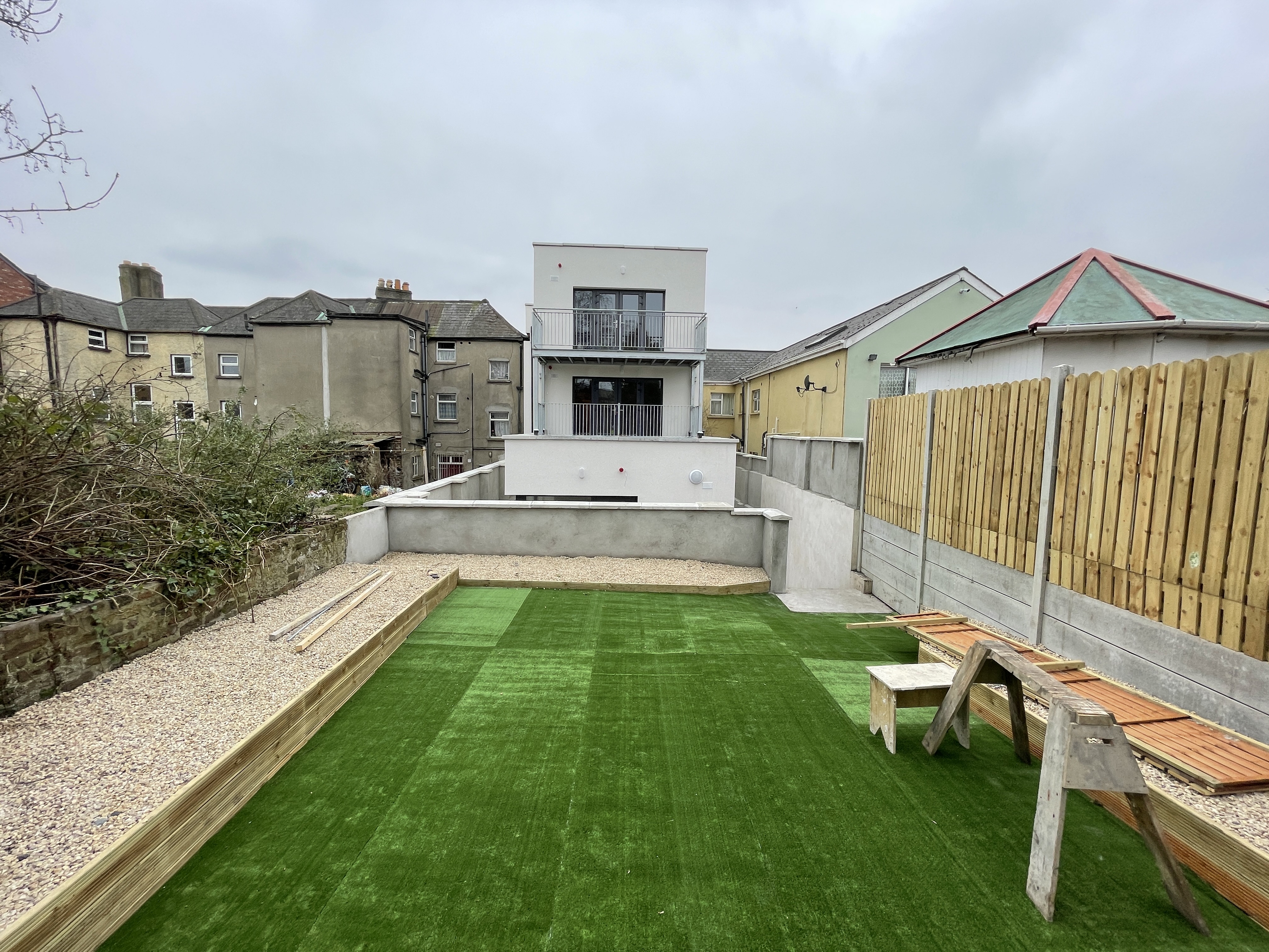 A rear yard with artificial grass and a picnic bench, behind a white-rendered apartment building.
