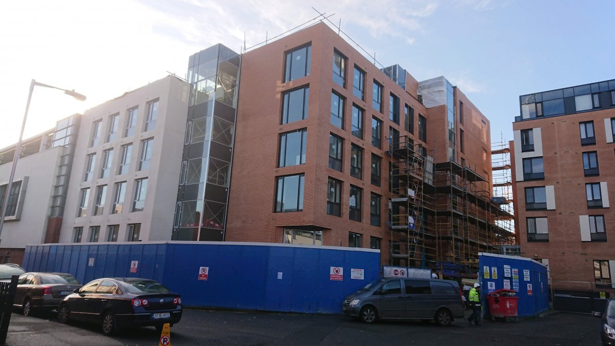 A red-brick student accommodation block under construction, with scaffolding and blue site hoarding along the footpath.